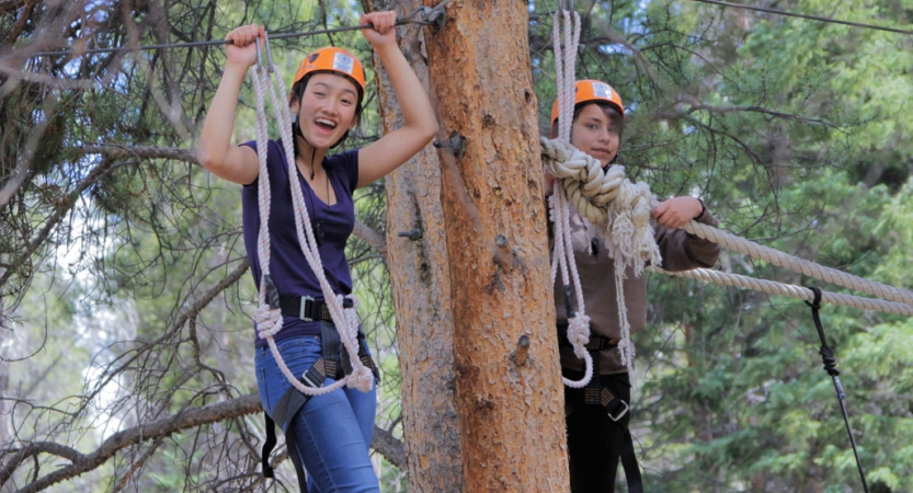 A student wearing safety gear is secured by ropes as they smile at the camera during a ropes course. 